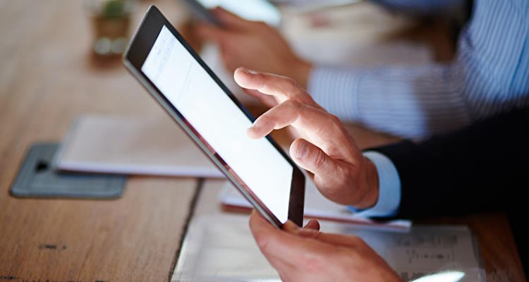 Person nutzt ein Tablet, während sie an einem Schreibtisch sitzt, mit unscharfen Händen einer weiteren Person im Hintergrund.