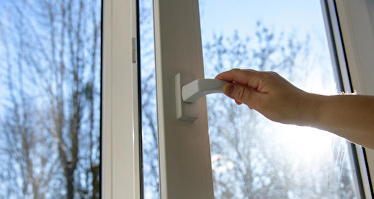 Hand öffnet ein Fenster, durch das Sonnenlicht und ein klarer Himmel sichtbar sind.
