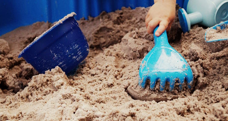 Sankasten mit Sandspielzeug. Eine Kinderhand benutzt eine hellblaue Spielharke.