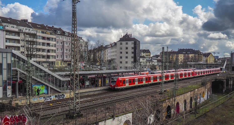 Im Hintergrund Wohngebäude. Im Vordergrund ein S-Bahnhof mit einfahrendem Zug. 