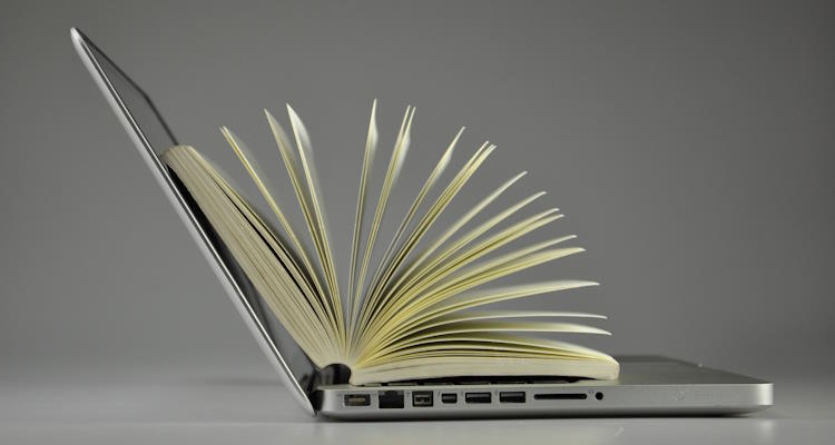 Ein aufgeklappter Laptop in der Seitenansicht. Auf der Tatstatur liegt ein aufgeschlagenes Buch.