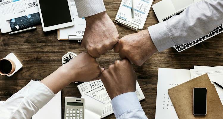 Symbolfoto: Schreibtisch mit unterschiedlichen Arbeitsmaterialien, in der Bildmitte berühren sich vier Menschen mit ausgestreckten Armen.