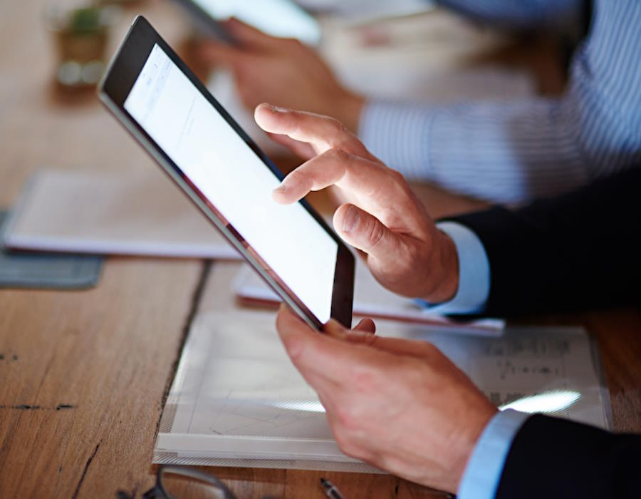 Person nutzt ein Tablet, während sie an einem Schreibtisch sitzt, mit unscharfen Händen einer weiteren Person im Hintergrund.