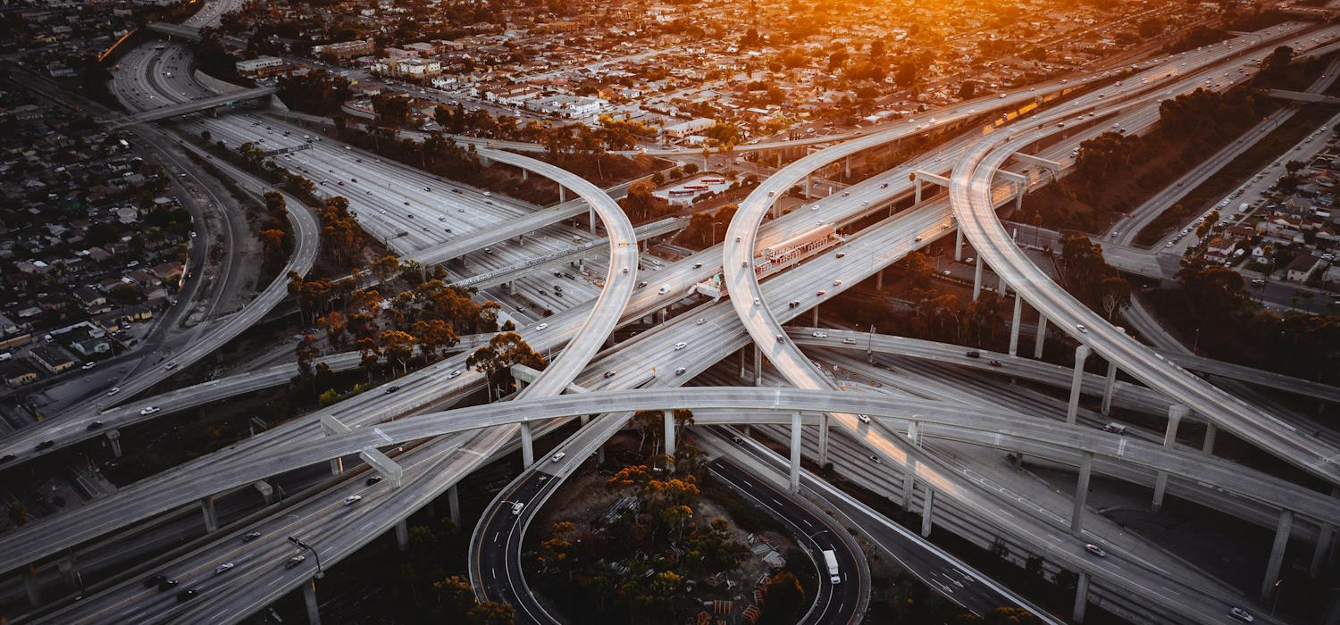 Symbolbild: Luftaufnahme eines großen, stark verzweigten Autobahnkreuz. Im Hintergrund ist eine Großstadt zu sehen. Die Sonne geht am Horizont auf. Symbolbild: Luftaufnahme eines großen, stark verzweigten Autobahnkreuz. Im Hintergrund ist eine Großstadt zu sehen. Die Sonne geht am Horizont auf.