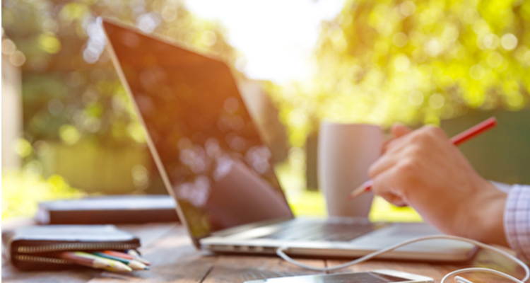 Person arbeitet im Freien am Laptop mit einer Tasse Kaffee und Stift in der Hand.