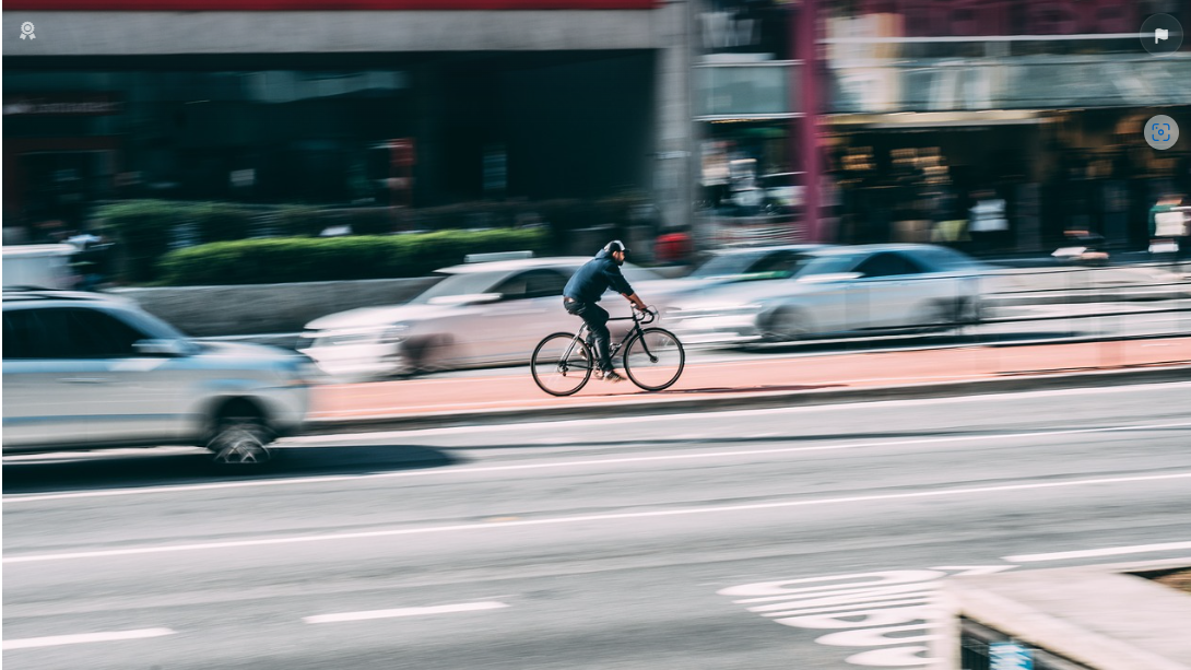 Das Bild zeigt Straßenverkehr mit Autos und einem Radfahrer. Während die Autos verwischt sind, liegt der Focus des Bildes auf dem Radfahrer.