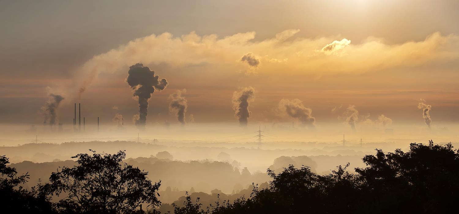Das Bild zeigt Landschaft bei Sonnenaufgang und Nebel. Im Vordergrund sind Wälder, im Hintergrund Industrieanlagen mit rauchenden Schornsteinen zu sehen.