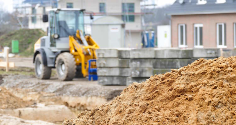 Symbolbild: Im Vordergrund ist ein großer Erdhaufen zu sehen. Im Hintergrund erkennt man einen gelben Bagger der auf einer noch nicht fertiggestellten Straße in einem Neubaugebiet Baumaterial transportiert.